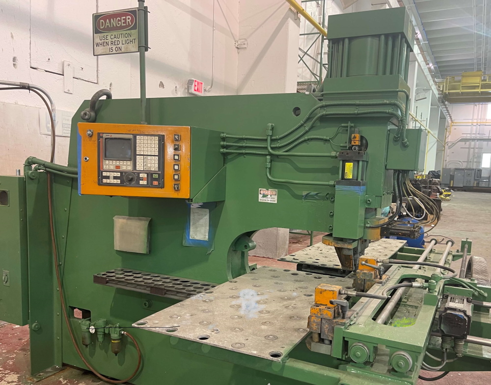 A green CNC plate drilling and punching machine with an orange control panel, drilled steel worktable and a Danger sign on the wall behind.