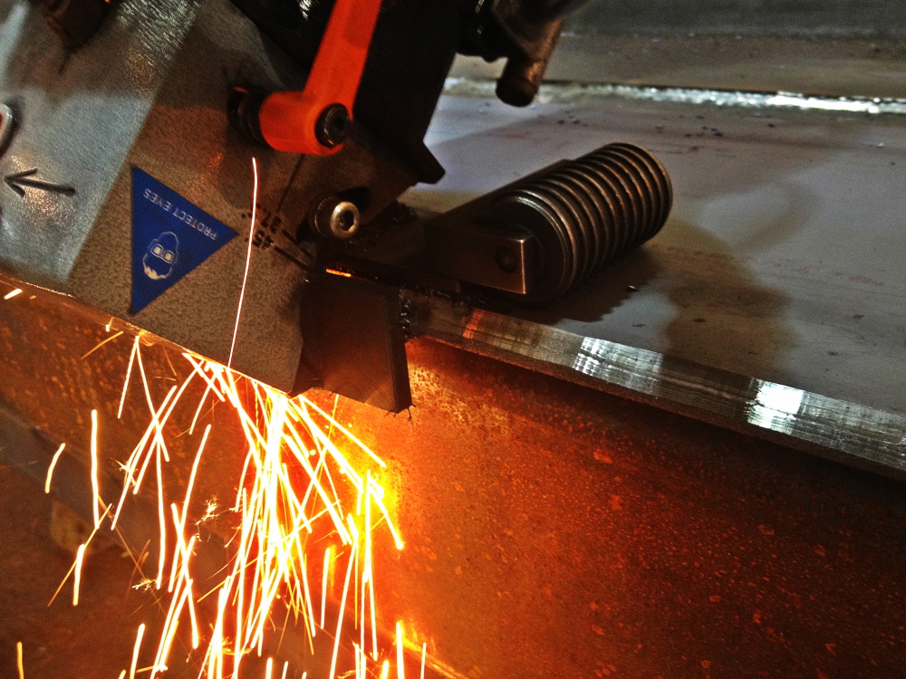 A band saw blade cuts into a steel section, throwing a bright spray of orange sparks with the freshly cut face shining alongside.