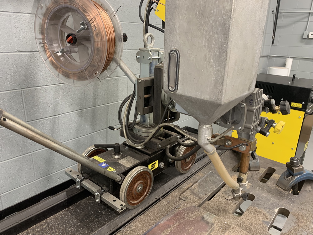 An ESAB submerged arc welding tractor sits on a track rail with a flux hopper and a spool of copper-coated wire, positioned over a plate joint.