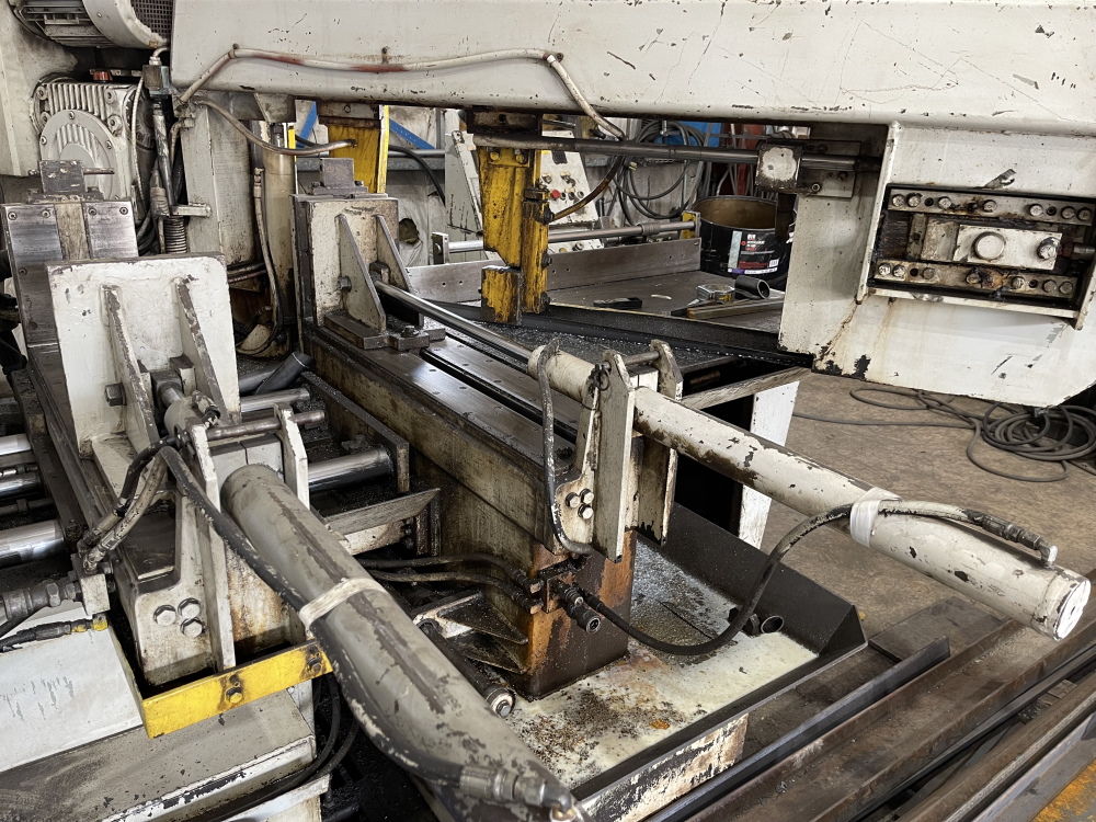 An automatic horizontal band saw in a fabrication shop, its hydraulic clamps and roller conveyor holding a steel section at the blade head.
