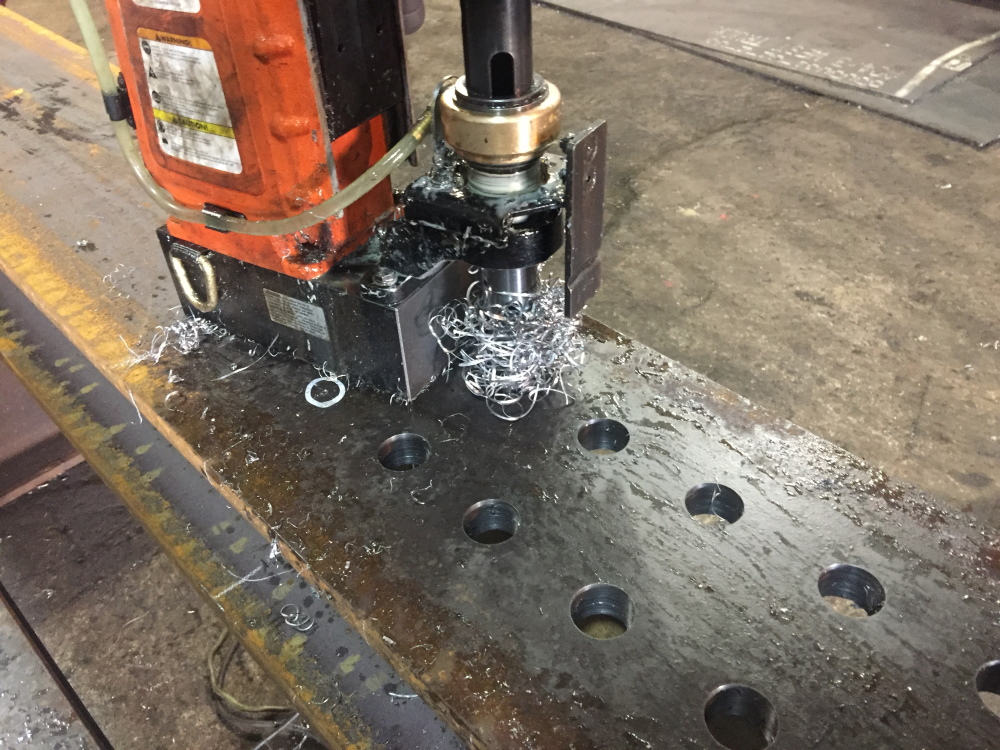 An orange magnetic drill press clamped to a steel beam flange, cutting a hole with curled metal chips beside an already drilled row of holes.