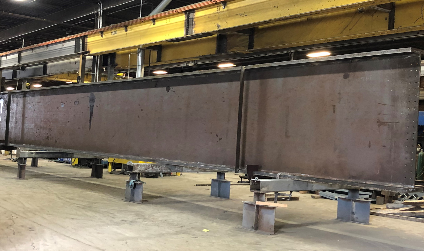 A long brown steel plate girder resting on short steel stands inside a fabrication shop, with a yellow overhead crane runway and shop lighting above.