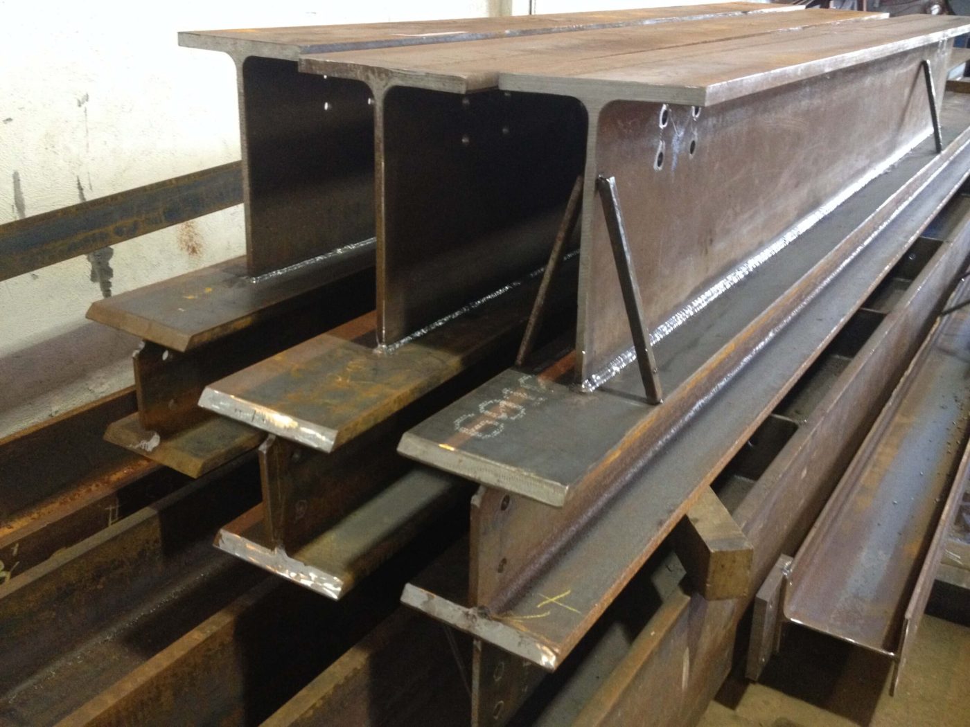 Built-up steel beams stacked in a shop, showing bright fillet welds where stiffeners and bearing plates are attached.
