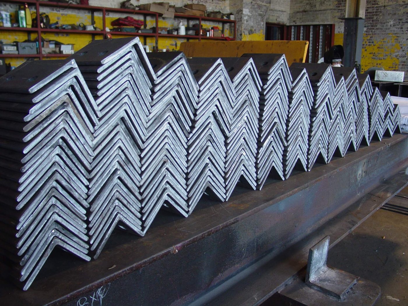 Rows of steel clip angles stacked in graded sizes along a beam in a fabrication shop, with tool shelving and a worker behind.