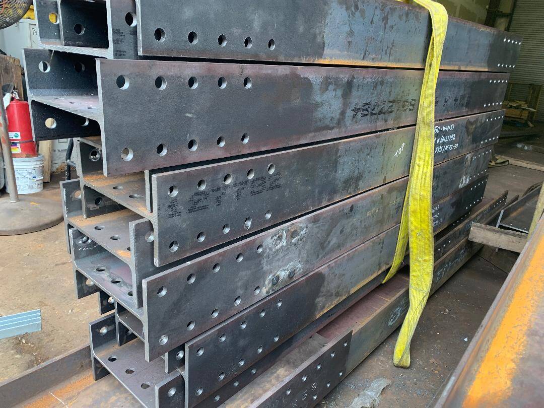 Several fabricated steel beams with rows of drilled bolt holes stacked one on top of another inside a fabrication shop, wrapped with a yellow lifting sling.