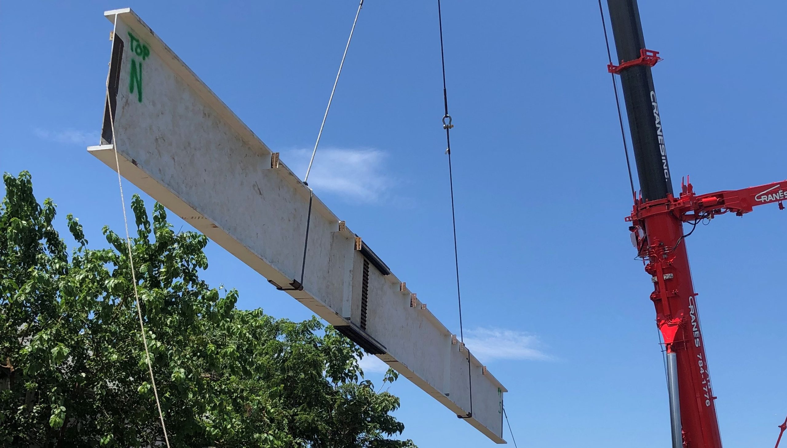 A long white-primed steel plate girder marked TOP in green paint hangs diagonally from crane chokers against a deep blue sky, with the boom of a red hydraulic truck crane at the right.