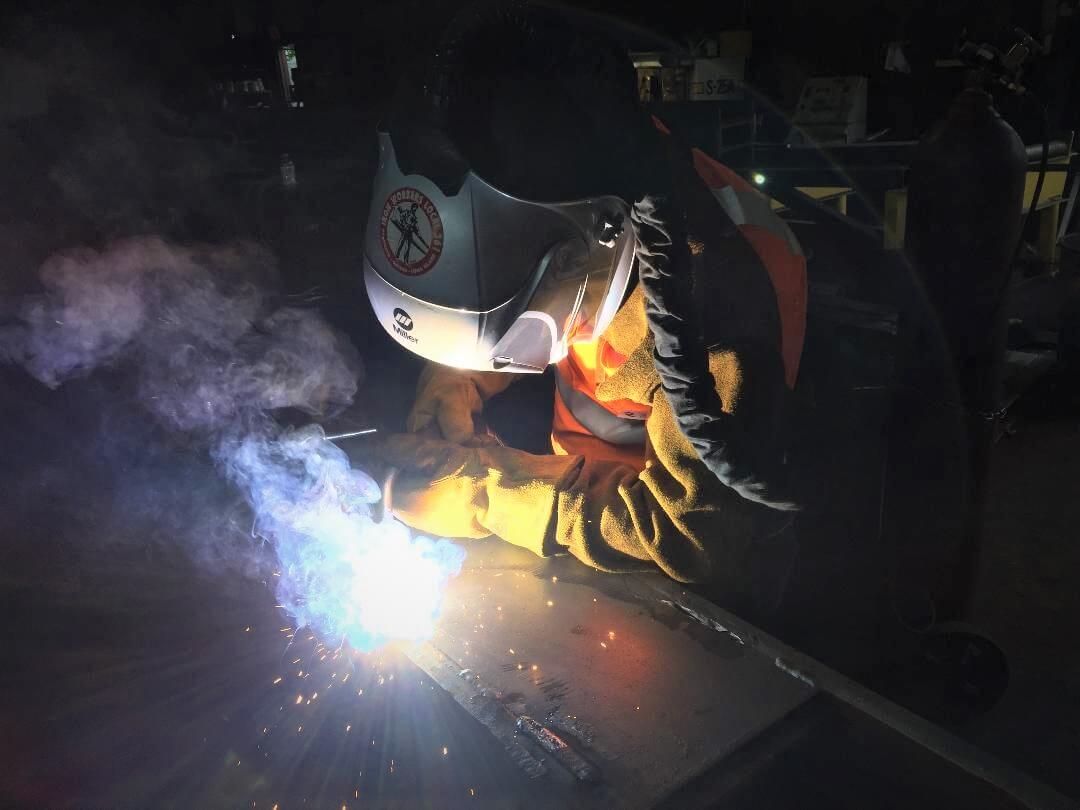 A welder in a hood and leather gloves striking a bright arc on a steel plate in a dark fabrication shop, with weld fume rising.
