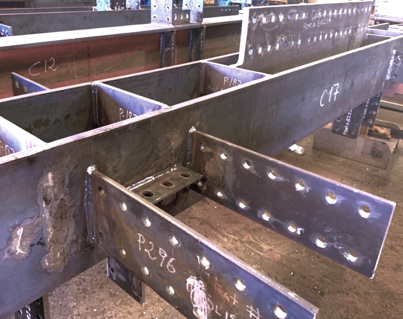 Welded built-up steel box sections and beams with internal stiffeners and drilled connection plates sit on a shop floor, marked with piece numbers in chalk.